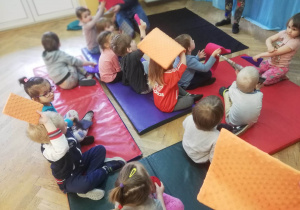 Dzieci siedzą na materacach trzymając w dłoniach poduszki w różnych kształtach.
