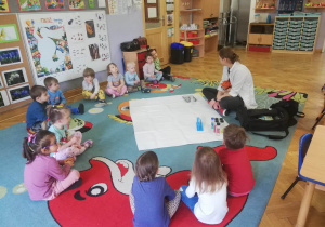 Dzieci siedzą w kole na dywanie, a prowadząca warsztaty chemiczne LABOLO opowiada im, czym będą zajmować się na zajęciach.