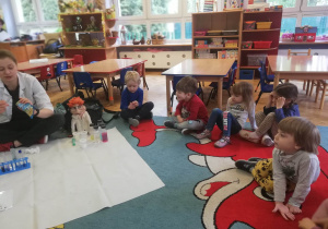 Dzieci siedzą na dywanie, a prowadząca warsztaty chemiczne LABOLO wykonuje doświadczenie, łącząc białka z różnymi cieczami.