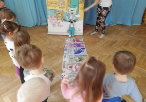 Dzieci stoją i oglądają różne nominały pieniędzy papierowych: 10 zł, 20zl, 50 zł, 100 zł, 200 zł i 500 zł.