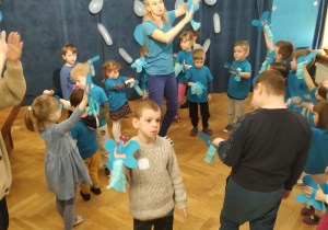 Dzieci ubrane na niebiesko tańczą z papierowymi niebieskimi motylkami na ręku.