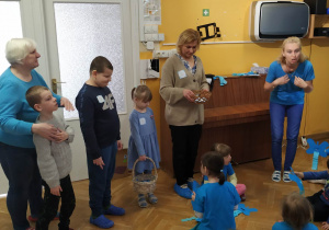 Dzieci ubrane na niebiesko siedzą na podłodze i śpiewają piosenkę z pokazywaniem, jako przywitanie dla przybyłych gości - dzieci z Przedszkole Specjalnego nr 208.
