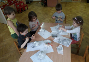 Dzieci siedzą przy stolikach i malują styropianowe zajączki szarą farbą.