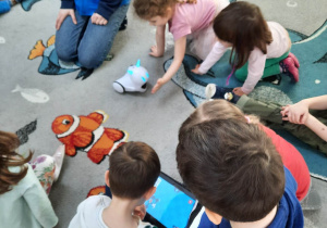 Dzieci siedzą na dywanie i programują na tablecie tor przejścia robota. Dzieci wybierają zwierzątko i rysują palcem jego tor przejścia po dywanie.