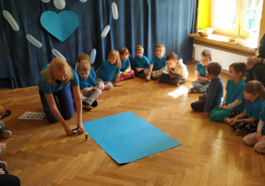 Dzieci siedzą w kole ubrane na niebiesko i obserwują, jak nauczycielka tłumaczy im ich zadanie - przyklejenie obrazków do plakatu.