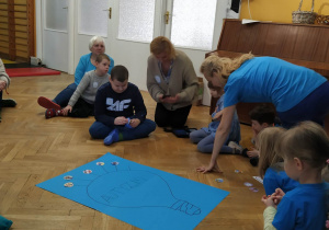 Dzieci siedzą na podłodze ubrane na niebiesko i przyklejają obrazki prezentujące fakty o autyzmie do plakatu.