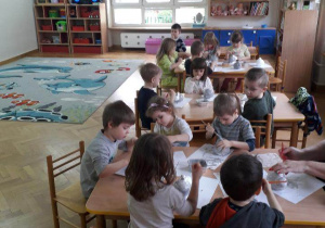 Dzieci siedzą przy stolikach i malują styropianowe zajączki szarą farbą.