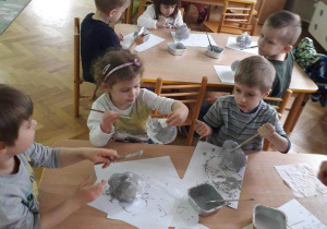 Dzieci siedzą przy stolikach i malują styropianowe zajączki szarą farbą.