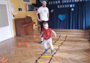 Chłopiec przeskakuje przez drabinkę z szarf ułożoną na podłodze podczas zajęć sportowych.