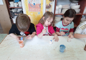 Chłopiec i dwie dziewczynki siedzą przy stoliku i malują specjalnymi farbami wcześniej ulepione własnoręcznie sówki ceramiczne.