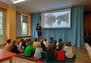 Dzieci siedzą na materacach i oglądają prezentację multimedialną nt. parków i przestrzeni zielonej w mieście.