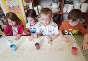 Dwóch chłopców i dwie dziewczynki siedzą przy stoliku i malują specjalnymi farbami wcześniej ulepione własnoręcznie sówki ceramiczne.