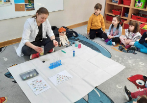 Dzieci siedzą na dywanie i obserwują, jak prowadząca warsztaty chemiczne LABOLO wykonuje doświadczenie chemiczne, dolewając ciecz do probówki.