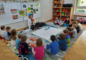Dzieci siedzą w półkolu na dywanie i witają się z prowadzącą warsztaty chemiczne LABOLO i kukiełką Profesorka Labolka.