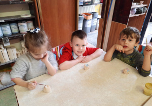 Dwóch chłopców i dziewczynka siedzą przy stoliku i malują specjalnymi farbami wcześniej ulepione własnoręcznie sówki ceramiczne.
