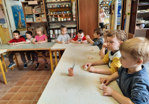 Dzieci siedzą przy stolikach i słuchają jak prowadząca warsztaty ceramiczne tłumaczy im czym będą się zajmowały.