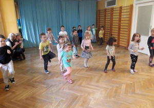 Dzieci stoją w rozsypce na sali gimnastycznej powtarzają ruchy za trenerką zumby podnosząc jedną nogę do góry.