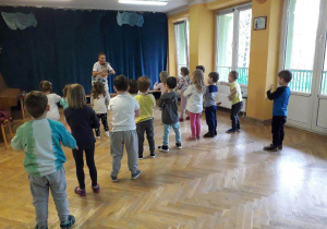 Dzieci stoją w rozsypce na sali gimnastycznej powtarzają ruchy za trenerką zumby kręcąc młynek rączkami.
