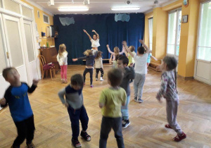 Dzieci stoją w rozsypce na sali gimnastycznej powtarzają ruchy za trenerką zumby podnosząc ręce do góry i machając nimi na boki.