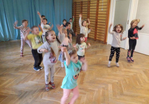 Dzieci stoją w rozsypce na sali gimnastycznej powtarzają ruchy za trenerką zumby podnosząc jedną nogę do góry. z wyciągniętymi do góry rękami.