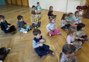 Dzieci siedzą w rozsypce na sali gimnastycznej powtarzają ruchy za trenerką zumby kręcąc młynek rączkami.