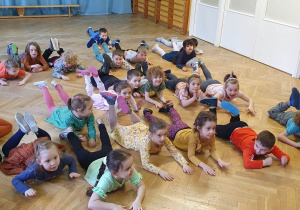 Dzieci leżą na podłodze na brzuchach w rozsypce na sali gimnastycznej powtarzając ruchy po trenerce zumby unosząc zgięte nogi do góry.