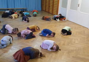 Dzieci leżą na podłodze w rozsypce na sali gimnastycznej w pozycji embrionalnej powtarzają ruchy za trenerką zumby.