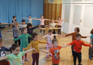 Dzieci stoją w rozsypce na sali gimnastycznej z rękami wyciągniętymi w bok, z dłońmi skierowanymi ku górze powtarzają ruchy po trenerce zumby.