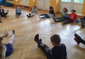 Dzieci siedzą w kole na podłodze podczas zajęć sportowych wykonując ćwiczenia unosząc jedną wyprostowaną nogę do góry.