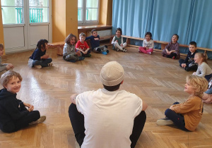 Dzieci siedzą w kole na podłodze podczas zajęć sportowych rozmawiając z prowadzącym zajęcia.