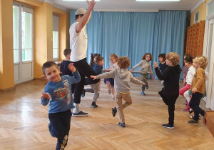 Dzieci ustawione w rozsypance na sali gimnastycznej wykonują ćwiczenia na równowagę, stojąc na jednej nodze podczas zajęć sportowych.