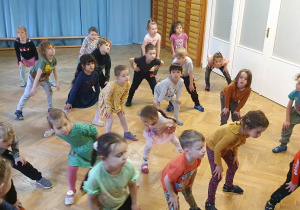 Dzieci stoją w rozsypce na sali gimnastycznej powtarzając ruchy po trenerce zumby stojąc z nogami ugiętymi w kolanach i trzymając ręce na nich.