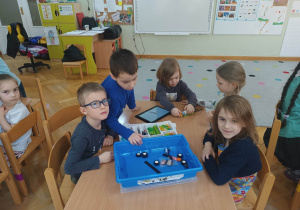 Dzieci siedzą przy stoliku i układają robota z klocków Lego wg instrukcji wyświetlanej na tablecie na zajęciach z robotyki i programowania.