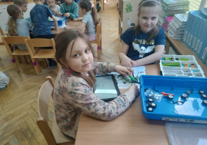 Dziewczynki siedzą przy stoliku i układają robota z klocków Lego wg instrukcji wyświetlanej na tablecie na zajęciach z robotyki i programowania.