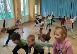 Dzieci stoją w rozsypce na sali gimnastycznej w rozkroku, pochylone do przodu z rękami uniesionymi za plecy do góry podczas zajęć z zumby.