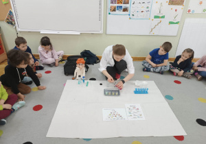 Dzieci siedzą w półkolu, a prowadząca warsztaty chemiczne LABOLO wykonuje doświadczenie chemiczne dodając różną ciecz do probówek.
