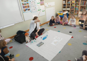 Dzieci siedzą w półkolu na dywanie, a prowadząca warsztaty chemiczne LABOLO pokazuje im probówkę z zabarwioną na fioletowo cieczą.