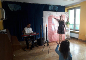 Prowadząca koncert interaktywny śpiewa piosenkę z prawą ręką wystawioną do bok, a Pan gra na keyboardzie.