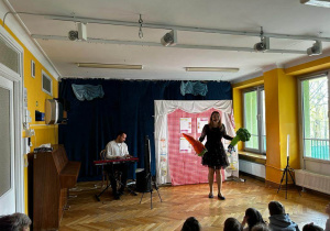 Prowadząca koncert interaktywny śpiewa piosenkę prezentując dzieciom maskotki marchewki i brokuła, a Pan gra na keyboardzie.
