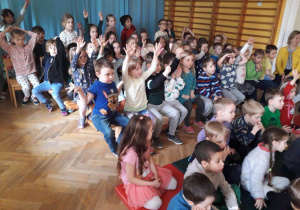 Dzieci siedzą na ławeczkach podczas koncertu interaktywnego podnoszą ręce do góry.