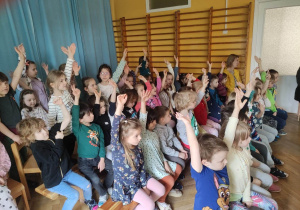 Dzieci siedzą na ławeczkach podczas koncertu interaktywnego podnoszą ręce do góry.
