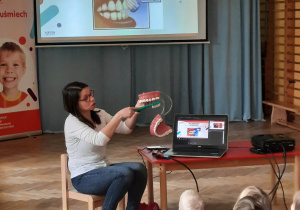 Dzieci siedzą na materacach i oglądają, jak prowadząca warsztaty stomatologiczne prezentuje im, jak należy szczotkować zęby, w tle wyświetlana jest prezentacja.