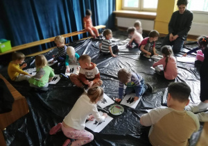 Dzieci siedzą na podłodze na folii i przyklejają czarno-białe obrazki do kartek i tworzą z nich pracę.