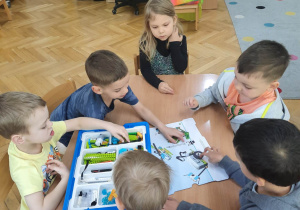 Dzieci siedzą przy stoliku i z pomocą papierowej instrukcji próbują złożyć robocika z kloców LEGO na warsztatach z robotyki.