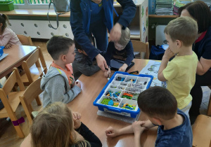 Dzieci siedzą przy stoliku, z pomocą prowadzącej warsztaty oraz instrukcji wyświetlanej na tablecie próbują zbudować robocika z klocków LEGO na warsztatach z robotyki.