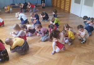 Dzieci ustawione w rozsypce powtarzają ruchy za trenerką zumby kucając i zakrywając głowy rękami.