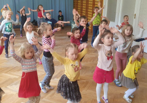 Dzieci ustawione w rozsypce powtarzają ruchy za trenerką zumby trzymają ręce machając nimi na boki w rytm muzyki.