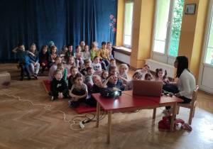 Dzieci siedzą na materacach i ławkach i słuchają prowadzącej warsztaty stomatologiczne.
