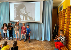 Kilkoro dzieci stoi pod ekranem rzutnika, na którym wyświetlana jest prezentacja nt. kina niemego i odgrywają scenkę, a reszta grupy ogląda.