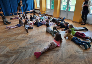 Dzieci leżą na podłodze na sali gimnastycznej podczas zajęć z zumby z nogami uniesionymi do góry i zgiętymi w kolanach obserwują trenerkę zumby.